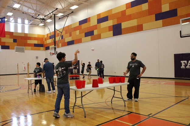 In the foreground, two young men play a cup-stacking game on a table in a gymnasium, while in the background, other people watch and a colorful wall mural adorns the far wall.