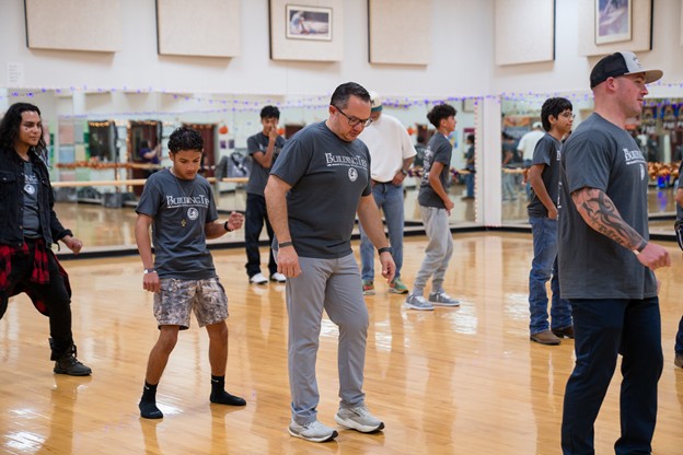 A group of young men and an instructor practice dance steps in a studio with mirrors and framed pictures on the walls.