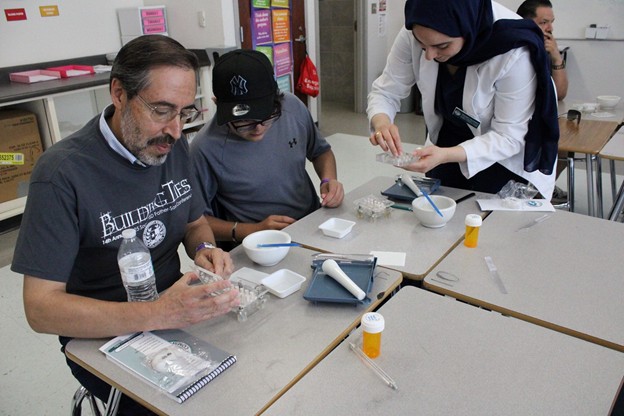 In the foreground, two men and a woman in a hijab are gathered around a table with small containers and tools, while the background shows a classroom setting with shelves and a whiteboard.