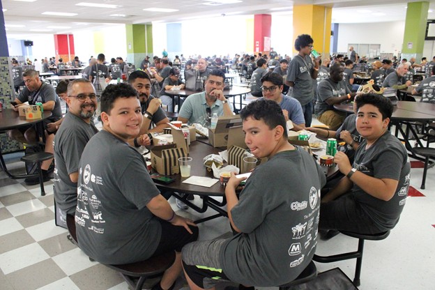 In the foreground, a group of young men in gray t-shirts sit around a table in a cafeteria, while in the background, many other people are seated at tables in a brightly lit, spacious room.