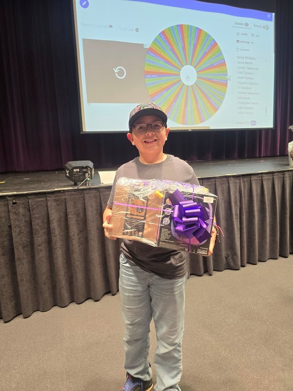 A smiling boy in a baseball cap and jeans holds a wrapped gift in the foreground, while a colorful prize wheel is displayed on a projector screen in the background.