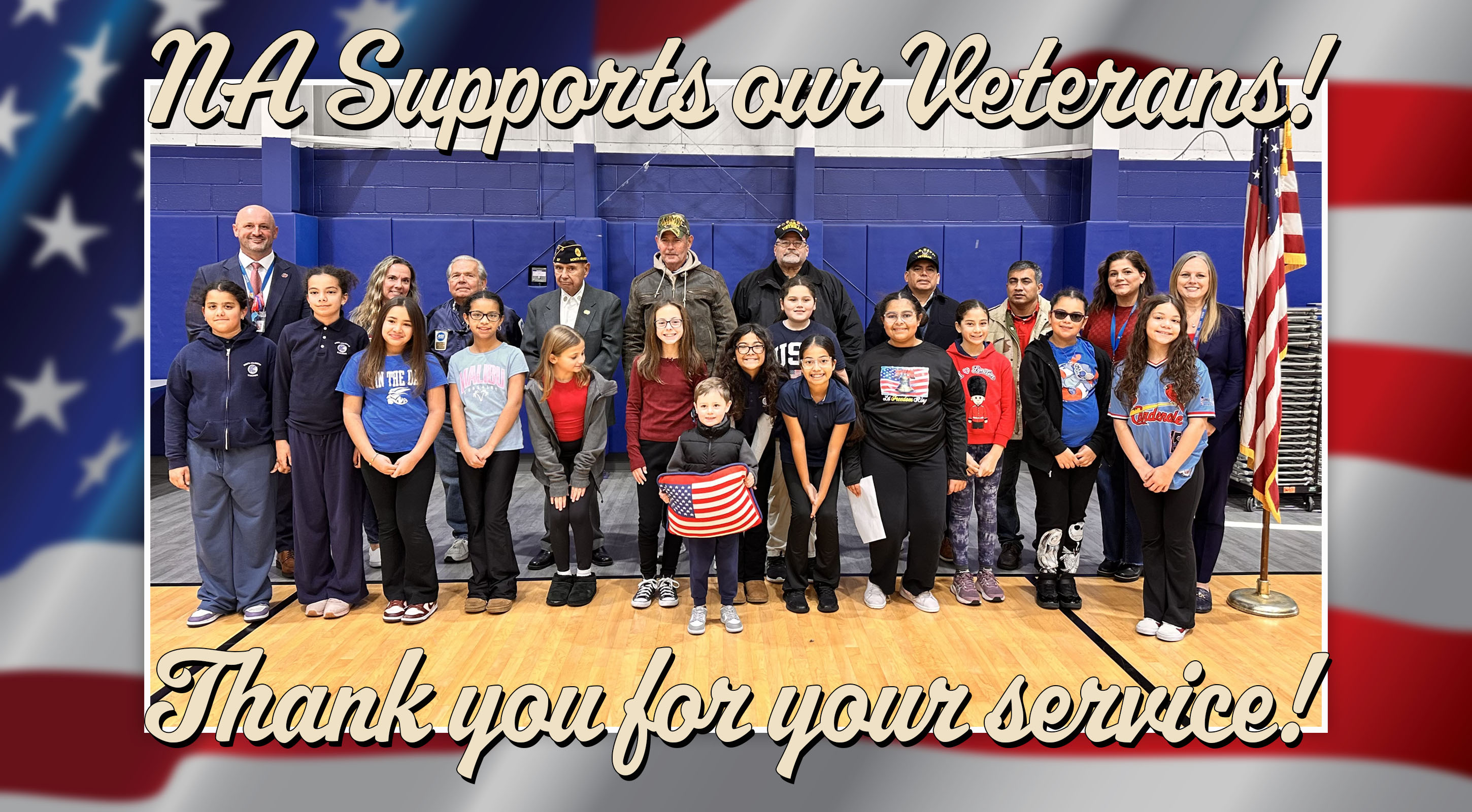 A group of students and veterans pose for a photo in front of a gymnasium backdrop, framed by an American flag.