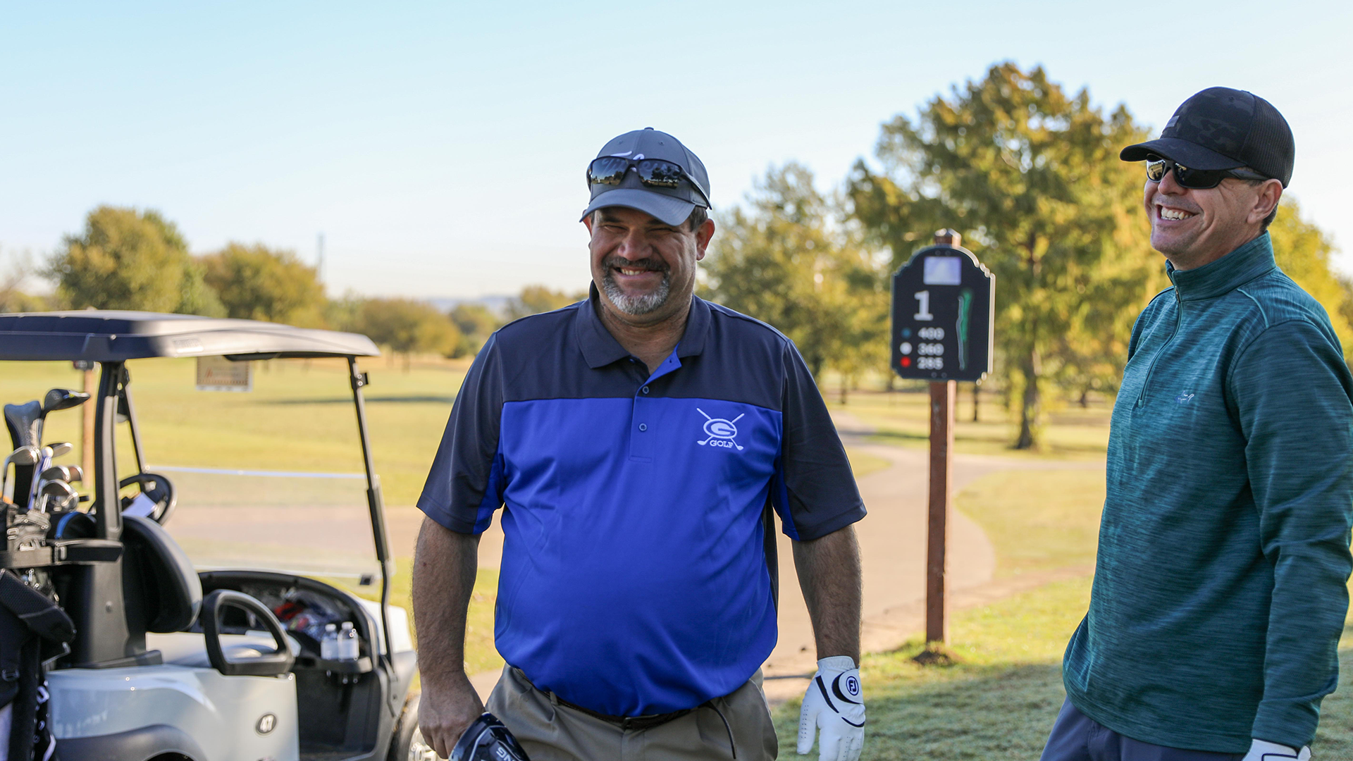 Two men in casual attire, one wearing a blue polo shirt and the other a green jacket, standing next to a golf cart on a grassy field with trees in the background.