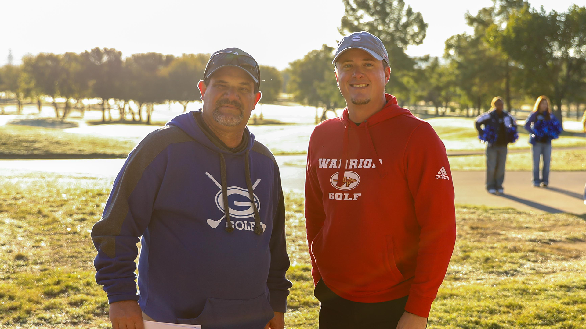 Two men wearing hoodies stand together in a grassy, wooded area on a sunny day.