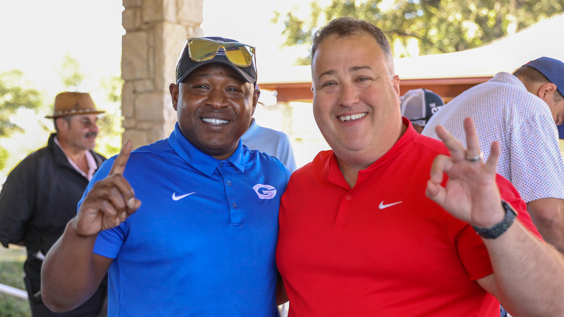 Two men wearing Nike shirts, one in blue and one in red, are smiling and posing together outdoors with a background of trees and buildings.
