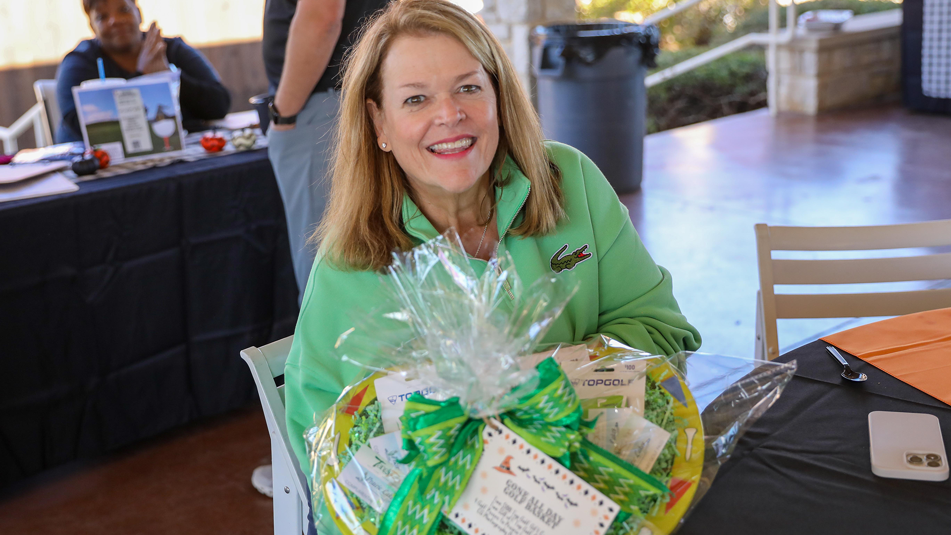 A smiling woman in a green jacket is holding a gift basket filled with various items, standing in what appears to be a conference or event setting with a black table and other items in the background.