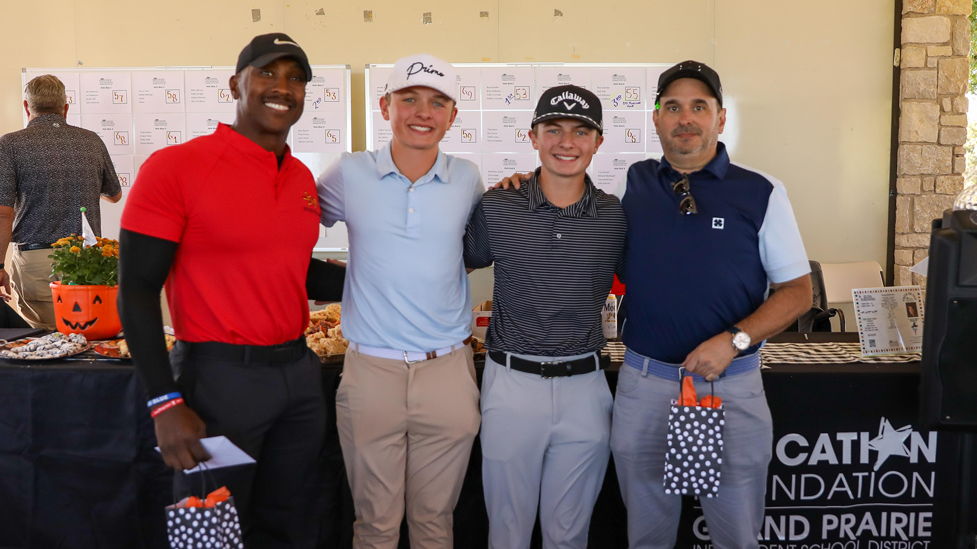The image shows four young men standing together, smiling and posing for a photograph. They are dressed in casual attire, including polo shirts, khakis, and baseball caps. The background appears to be an office or workplace setting, with a bulletin board and other office-related items visible.
