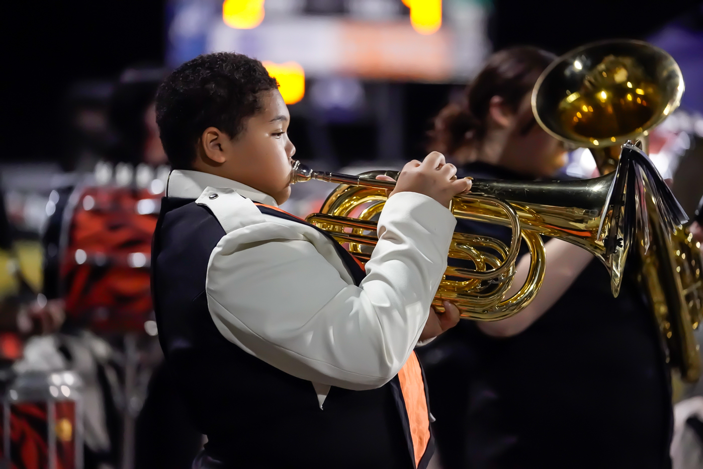 A young person in a band uniform plays a brass instrument outdoors at night, with other band members visible in the blurred background.