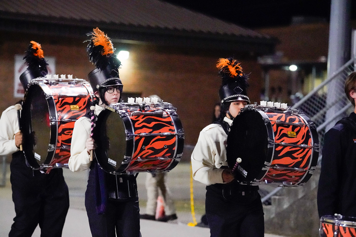 A high school marching band in black and white uniforms with orange accents performs outside. Several members play clarinets and saxophones, wearing hats with black and orange plumes. A brick building is in the background.