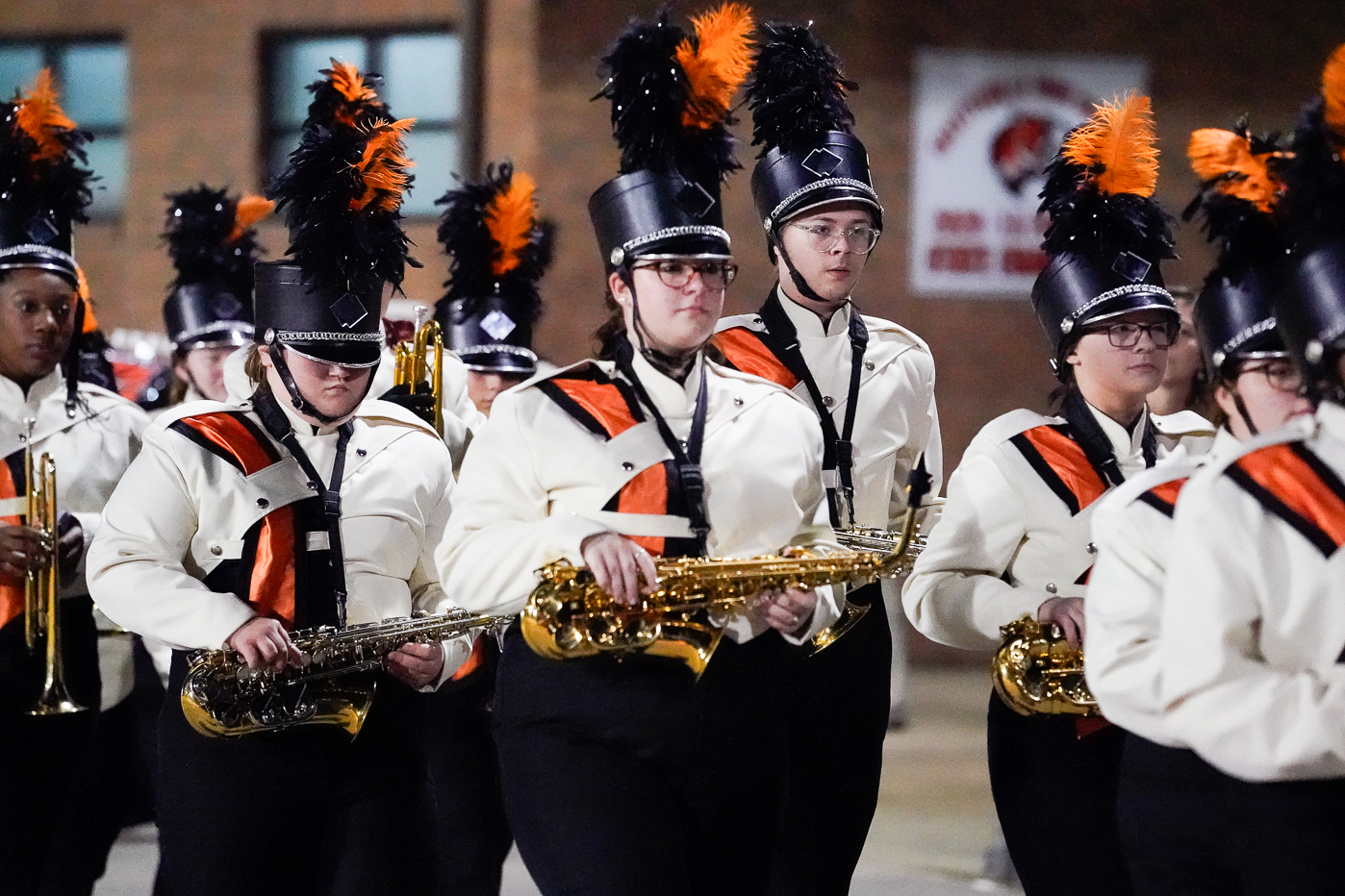 A marching band dressed in white uniforms with black and orange accents, wearing tall hats with orange and black plumes, marches in formation while playing saxophones and trumpets.