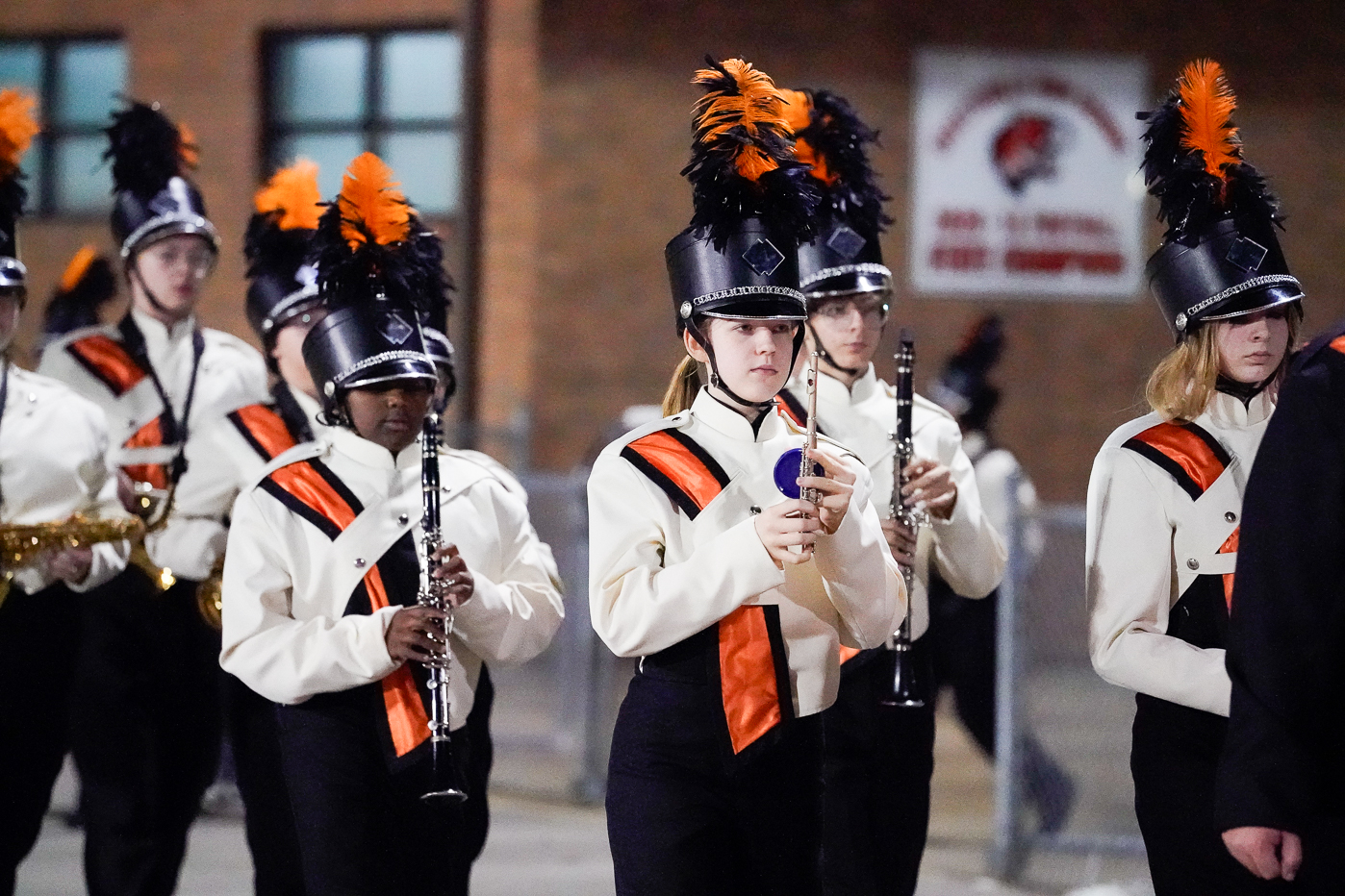 A high school marching band in black and white uniforms with orange accents performs outside. Several members play clarinets and saxophones, wearing hats with black and orange plumes. A brick building is in the background.