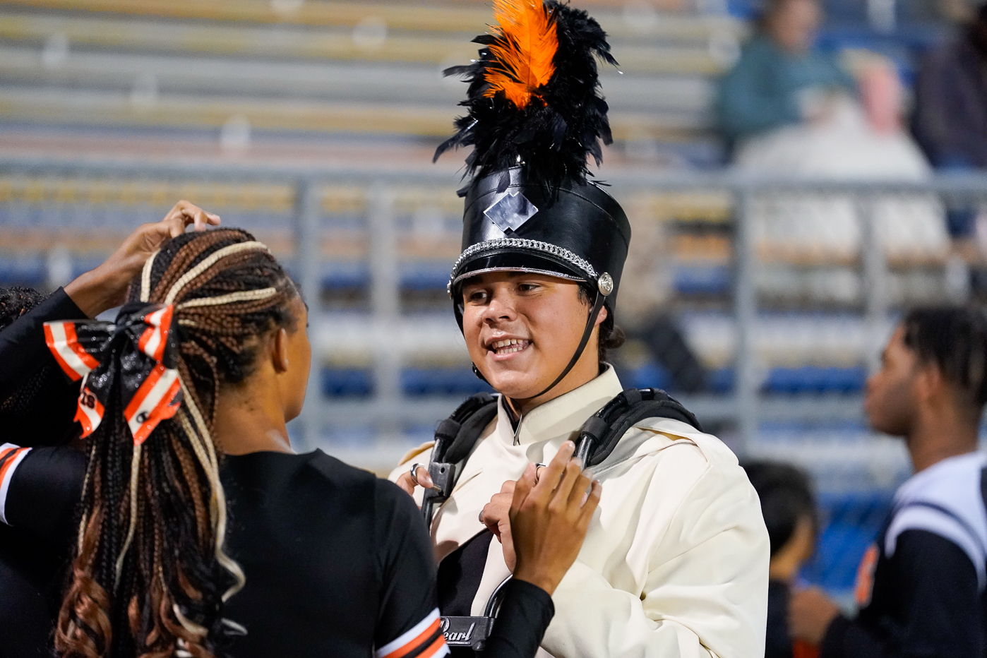 A marching band member in a white uniform and tall black hat with an orange and black plume smiles while talking to cheerleaders wearing black outfits with orange and white bows.