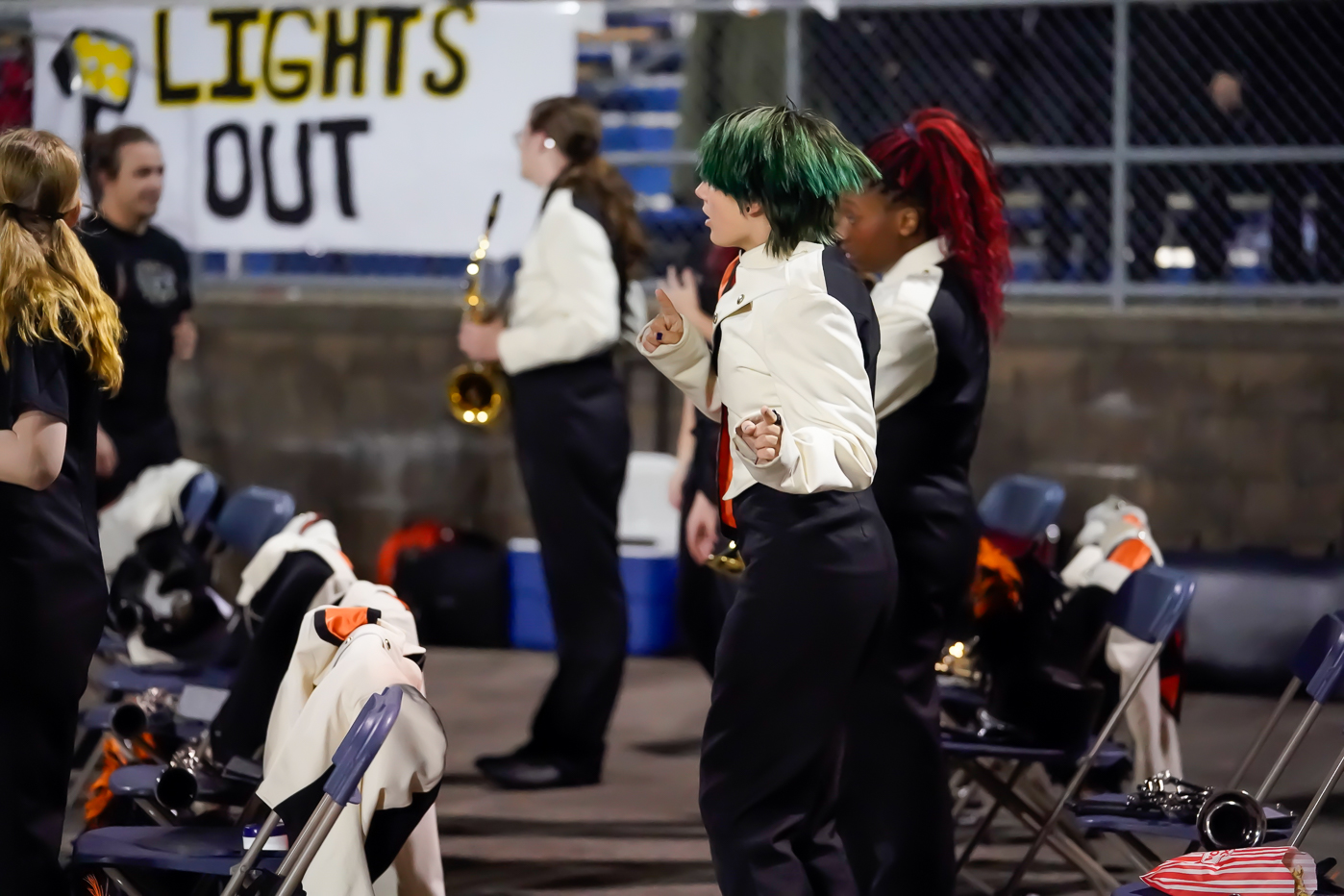 A person with green hair in a band uniform stands and gestures energetically near folding chairs, with other band members and a Lights Out sign visible in the background.