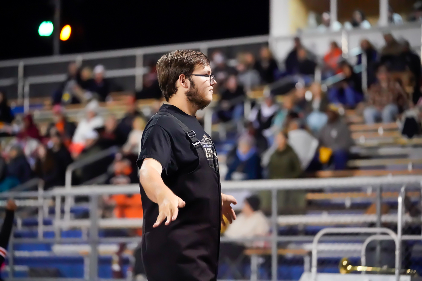 A young man wearing glasses and a black shirt stands with arms slightly raised at a stadium, as blurred spectators sit in bleachers behind him.