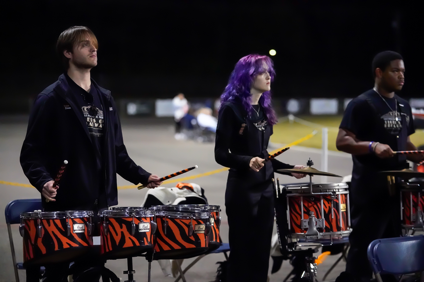 Three percussionists play drums outdoors at night. The drums have orange and black tiger stripes. The middle person has long purple hair. They are focused, wearing black shirts, and standing behind blue chairs.