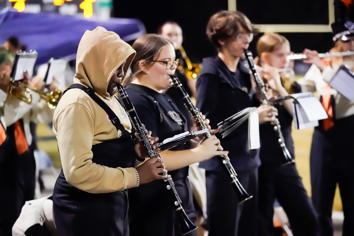 A group of band members play clarinets during a nighttime outdoor performance. They are focused on their music, wearing uniforms, and standing in rows with blurred musicians visible in the background.