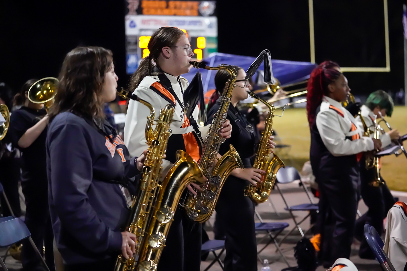A high school marching band plays saxophones and brass instruments while standing on a sideline at night, wearing black and white uniforms with orange sashes, with a scoreboard visible in the background.
