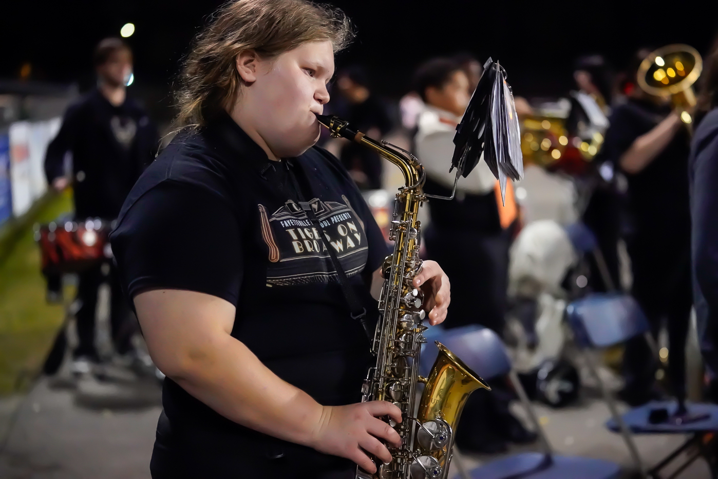 A person in a black shirt plays a saxophone outdoors at night, surrounded by other musicians and empty chairs, as part of a band or musical group.