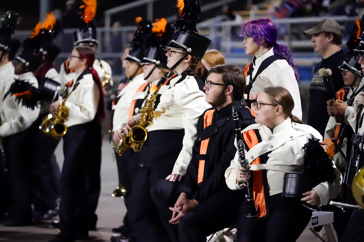 A group of marching band members in white and black uniforms with orange accents sit and stand together, holding instruments such as saxophones and clarinets, at an outdoor event at night.