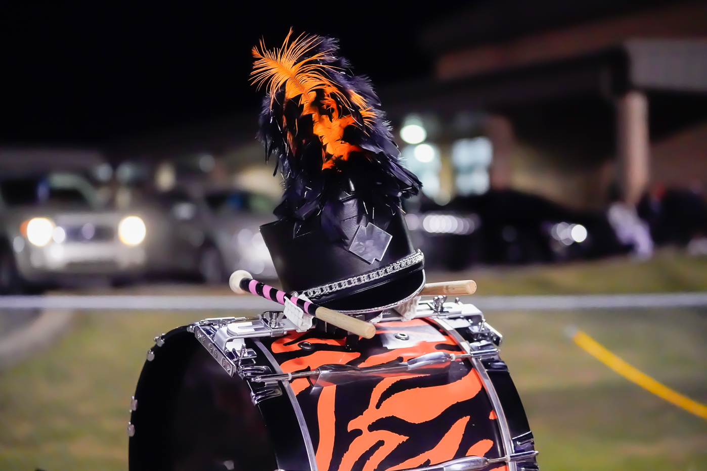 A marching band drum with an orange and black tiger-striped cover, a decorated hat with black and orange feathers, and a pair of drumsticks resting on top. The background is blurred with cars and a building.