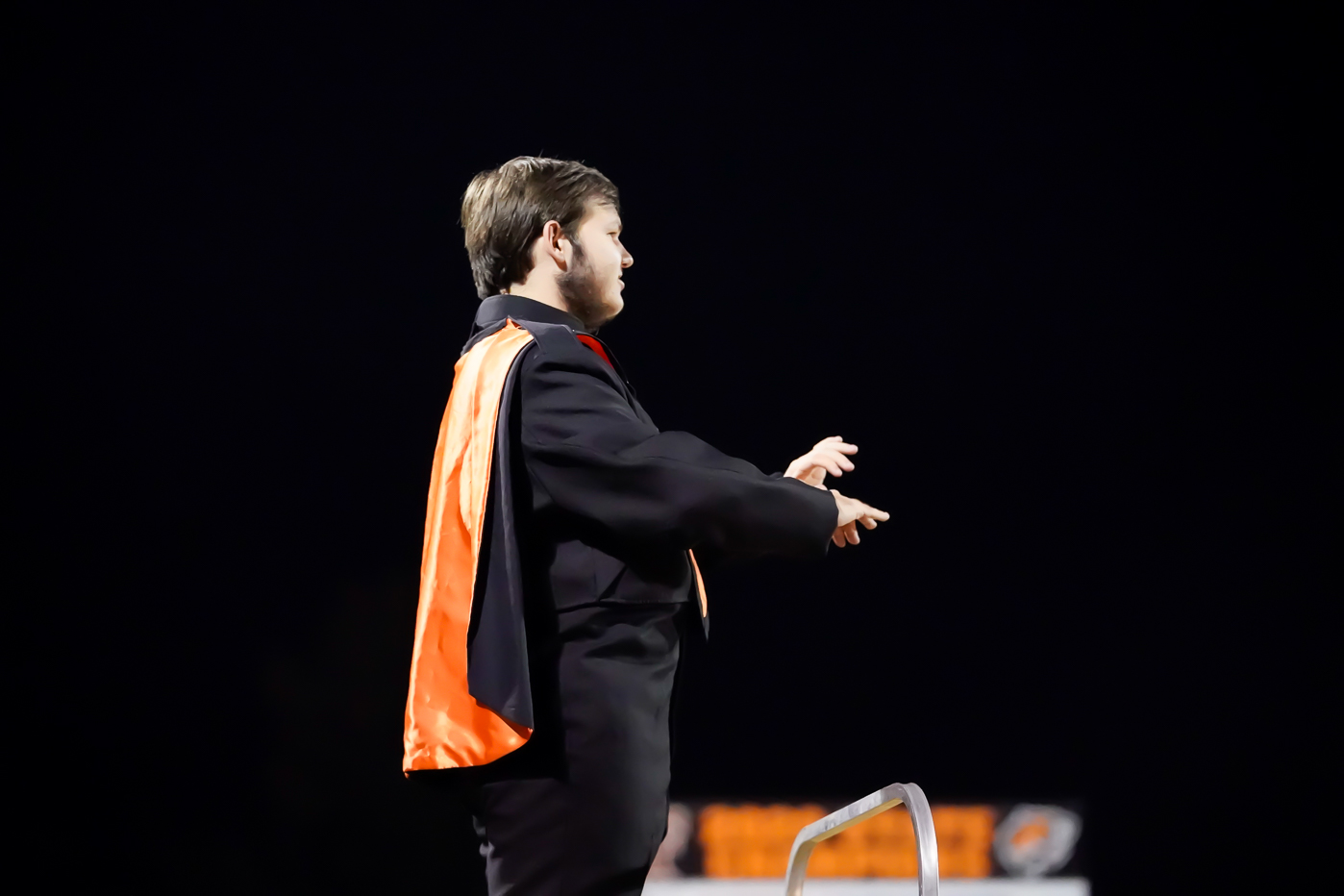 A young person in a black suit with an orange sash stands on a platform, conducting with both hands raised against a dark night sky.