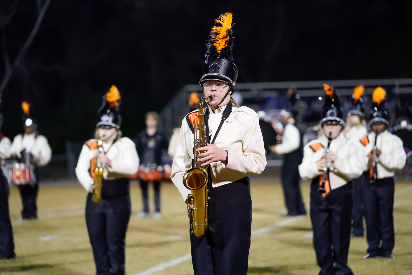 A marching band performs at night on a field. The focus is on a saxophone player wearing a black and orange plume on their helmet, with other band members and instruments in the background.