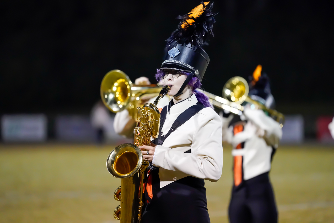 A marching band member wearing a black hat with an orange feather plays a saxophone on a field, with another band member playing a brass instrument in the background.