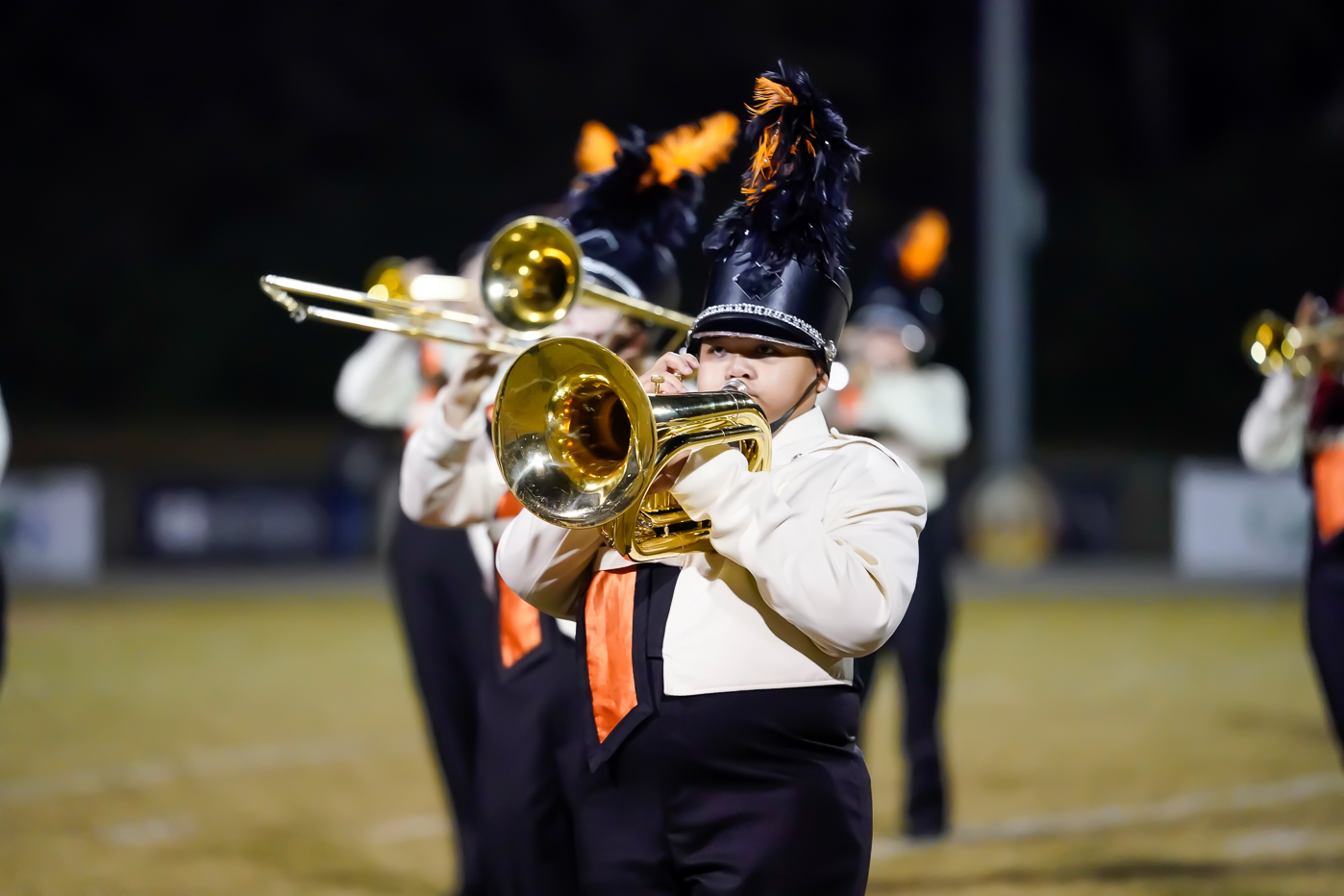 A marching band member in uniform with a feathered hat plays a brass instrument, with other band members in matching attire blurred in the background on a field at night.