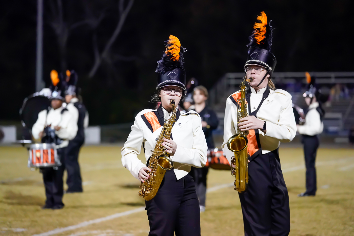 Two marching band members in white uniforms with orange and black plumes play saxophones on a football field, with other band members and instruments visible in the background.