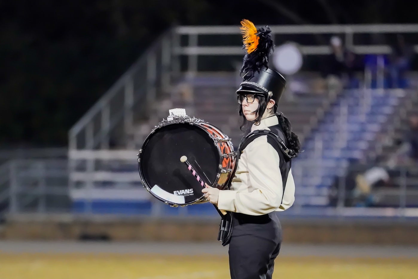 A marching band member in uniform and a plumed hat stands with a large bass drum during a performance on a field, with empty bleachers visible in the background.