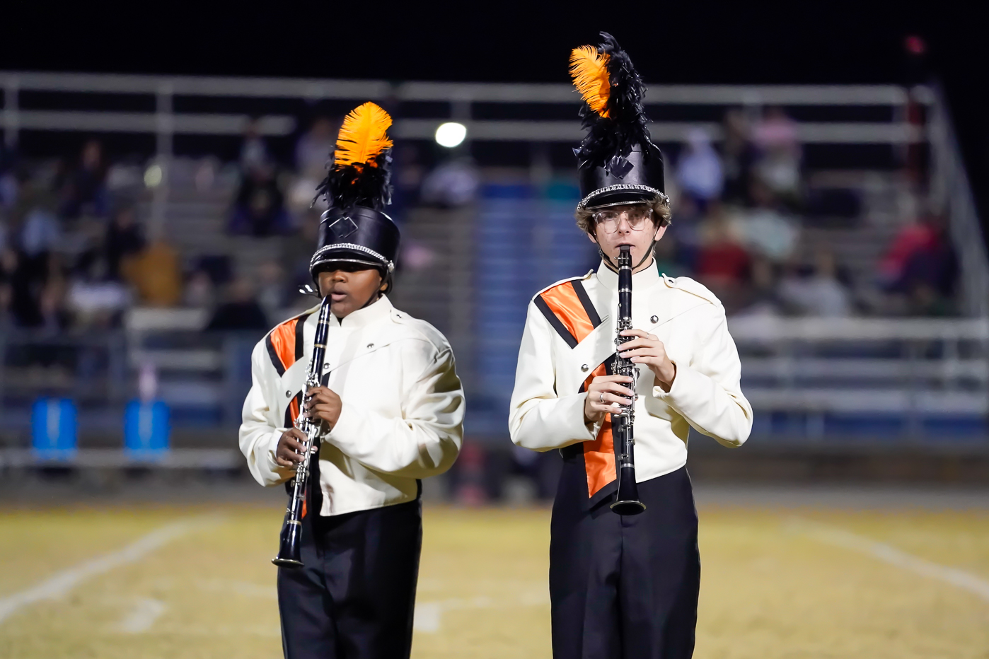 Two marching band members in white uniforms with orange and black sashes and feathered hats play clarinets on a field. Blurred stadium seating and spectators are visible in the background at night.