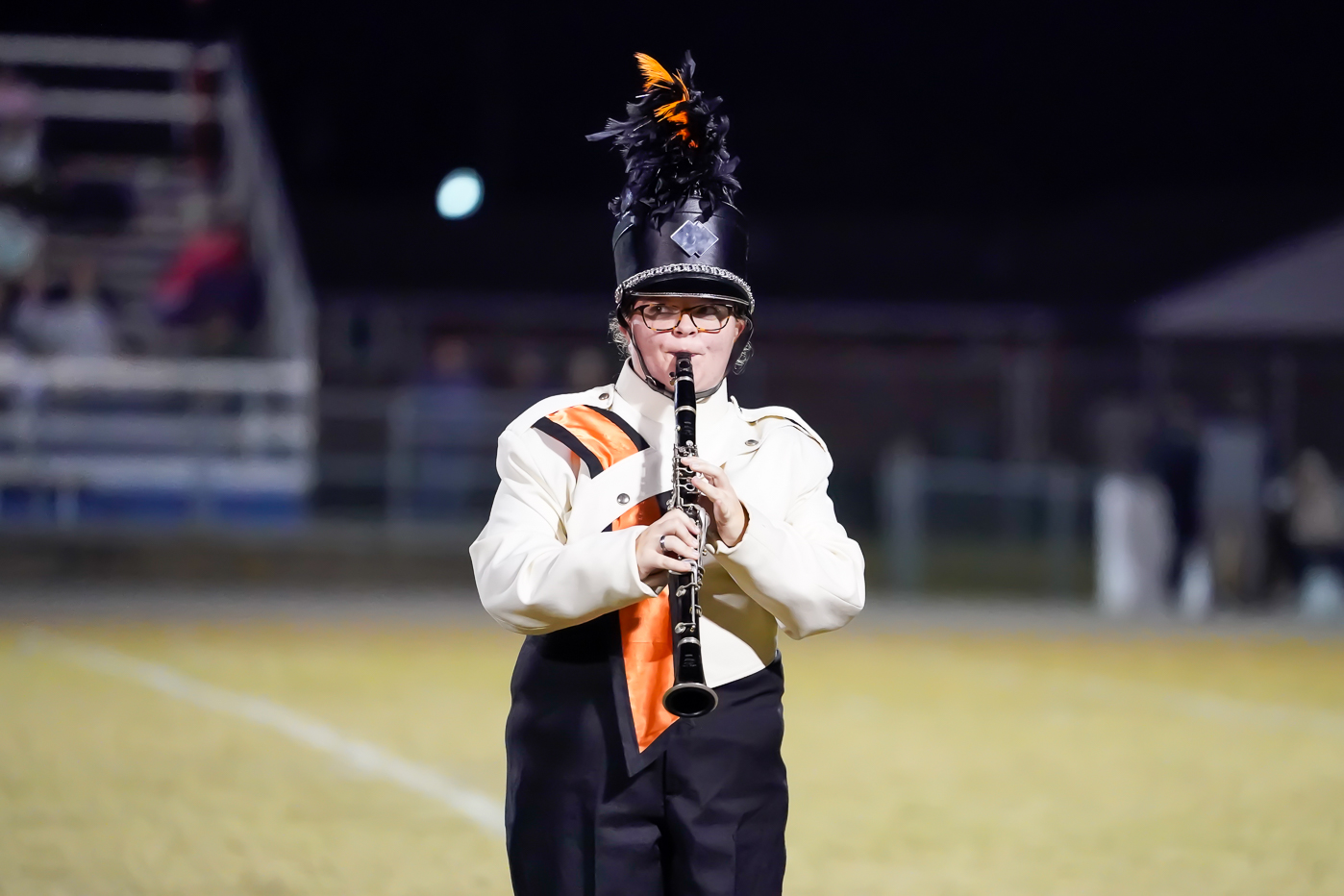 A marching band member in a uniform with orange and black stripes and a tall plume on their hat plays a clarinet on a football field at night. Bleachers and blurred spectators are in the background.
