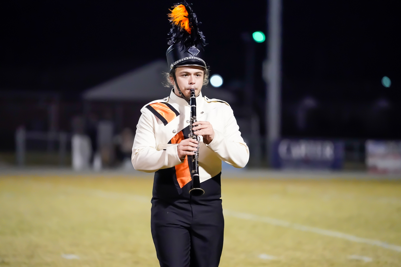 A marching band member in uniform plays a clarinet on a field at night. The uniform is white and black with orange accents, and the hat has black and orange plumes. The background is blurred.