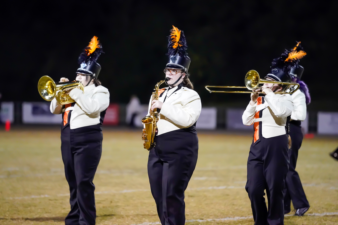 Three marching band members in white shirts, black pants, and tall hats with orange plumes perform with brass and woodwind instruments on a field at night.