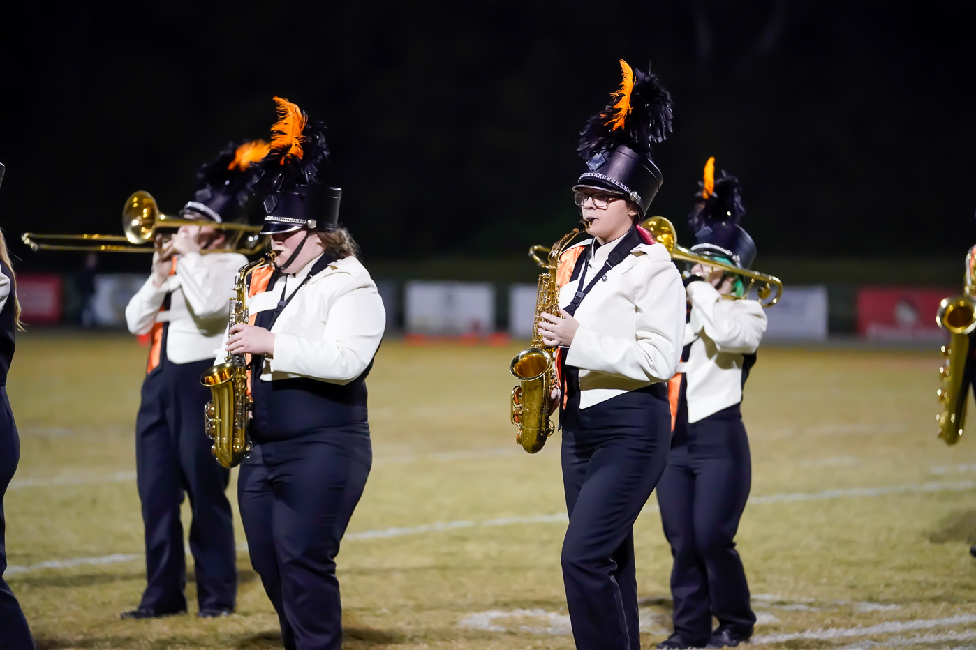 A marching band performs on a field at night, with members wearing black and white uniforms, orange ties, and hats with black and orange plumes while playing brass and woodwind instruments.