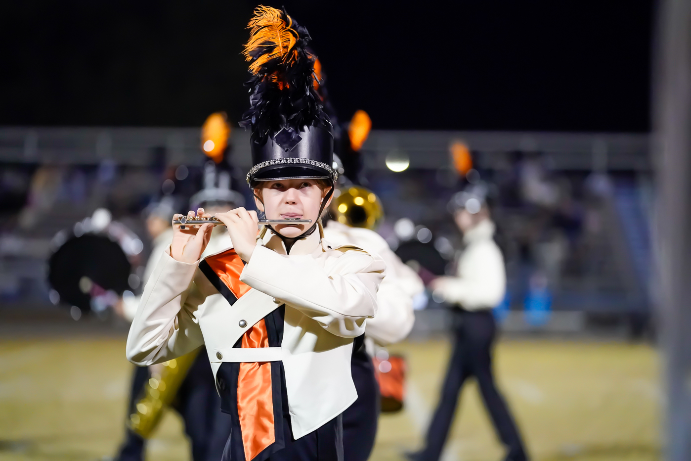 A marching band member in a white and black uniform with orange accents plays the flute on a field at night, with other band members blurred in the background.