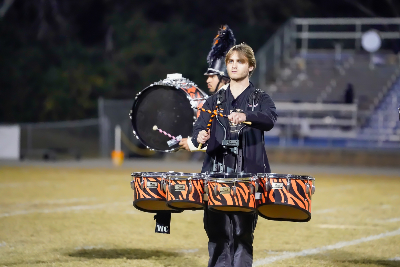 A marching band member plays tiger-striped tenor drums on a football field at night, with bleachers and another drummer in the background.