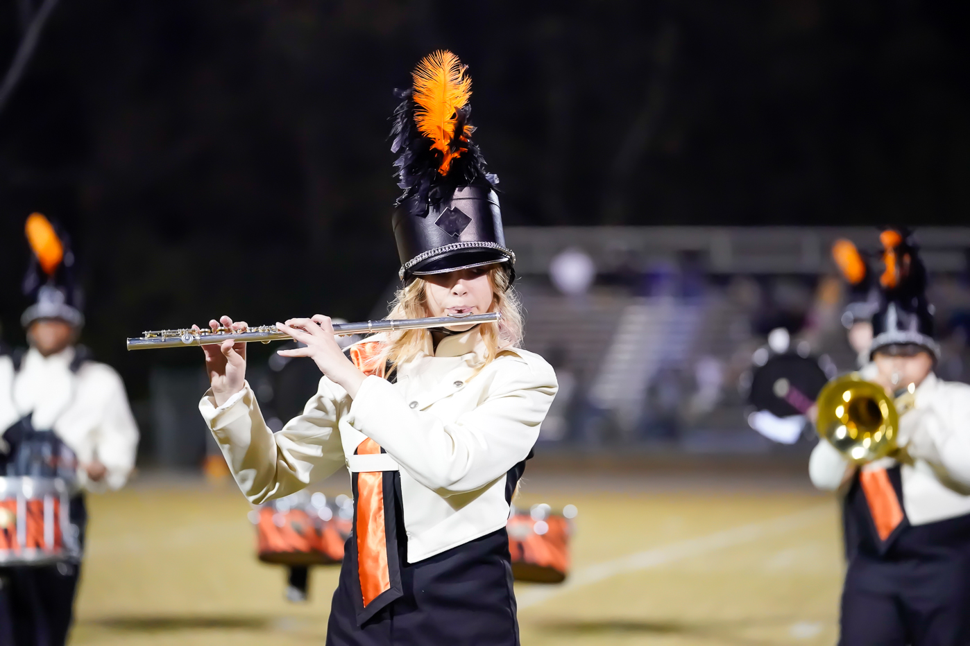 A marching band member in a white and black uniform with orange accents and a feathered hat plays the flute on a field, with other band members and bleachers visible in the background.