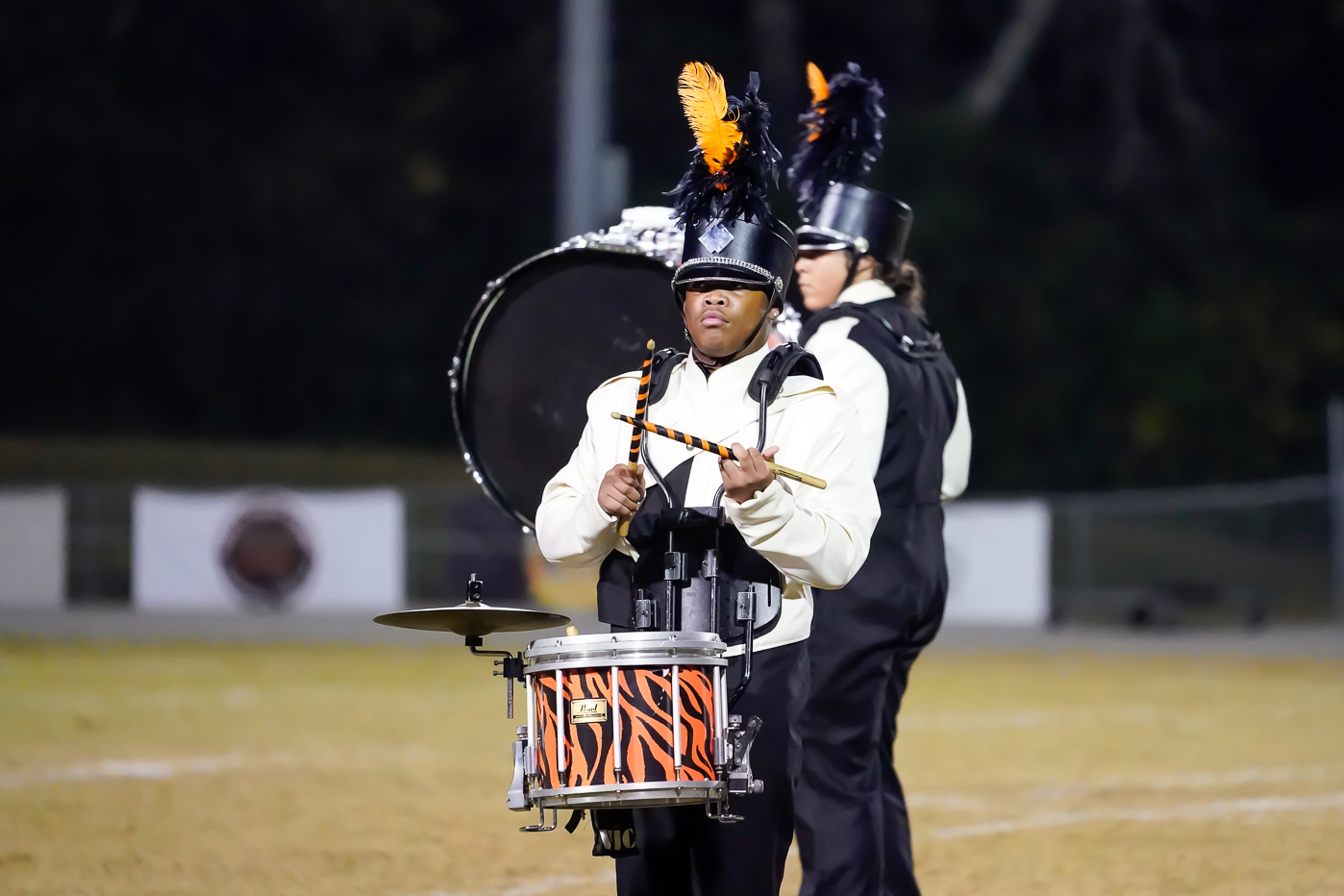 A marching band member in a white and black uniform with orange feathered plumes plays a snare drum with an orange-striped design. Another member, partially visible, stands behind with a large drum. They are on a field at night.