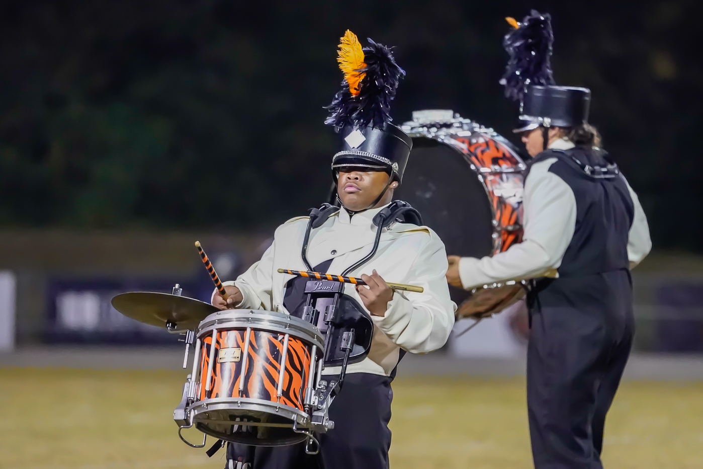 A marching band drummer in uniform plays a snare drum with orange and black stripes, wearing a tall hat adorned with black and orange feathers. Another band member with a bass drum stands behind.