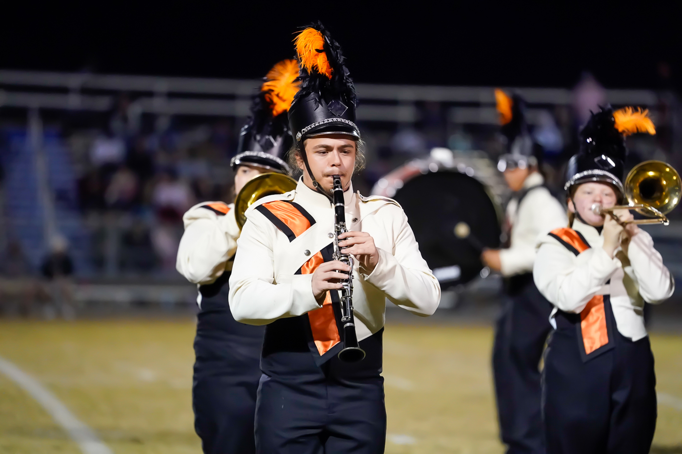 A marching band member in uniform plays a clarinet on a field at night. The uniform is white and black with orange accents, and the hat has black and orange plumes. The background is blurred.
