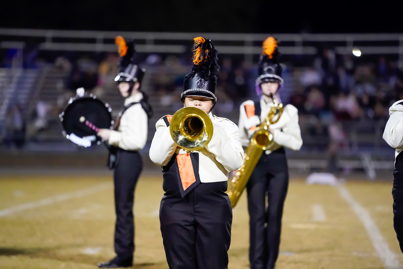 A marching band member in uniform plays a brass instrument on a football field, with two other uniformed band members holding instruments in the blurred background. Bleachers with spectators are visible behind them.