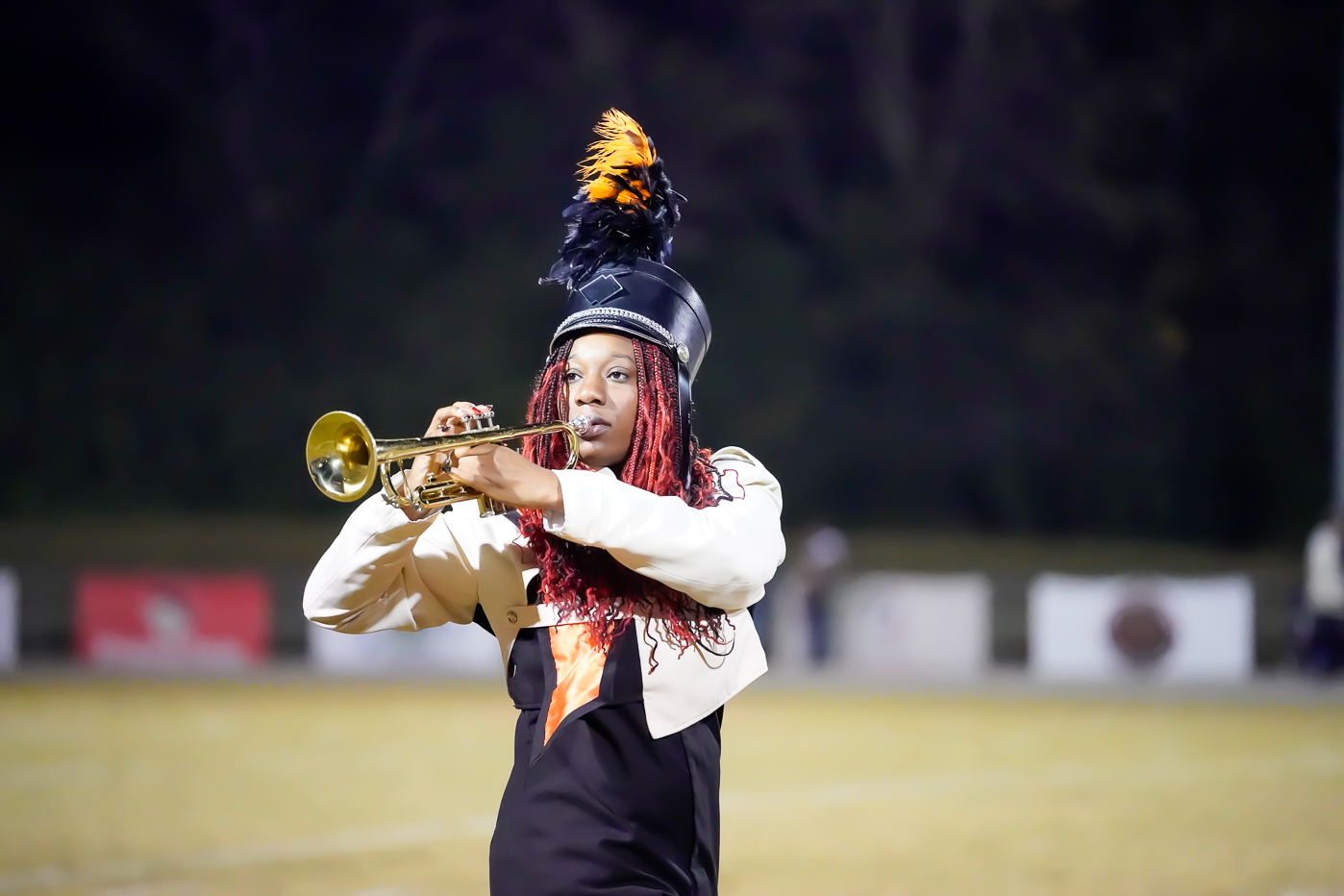A marching band member wearing a black and gold uniform with a feathered hat plays a trumpet outdoors on a field at night.