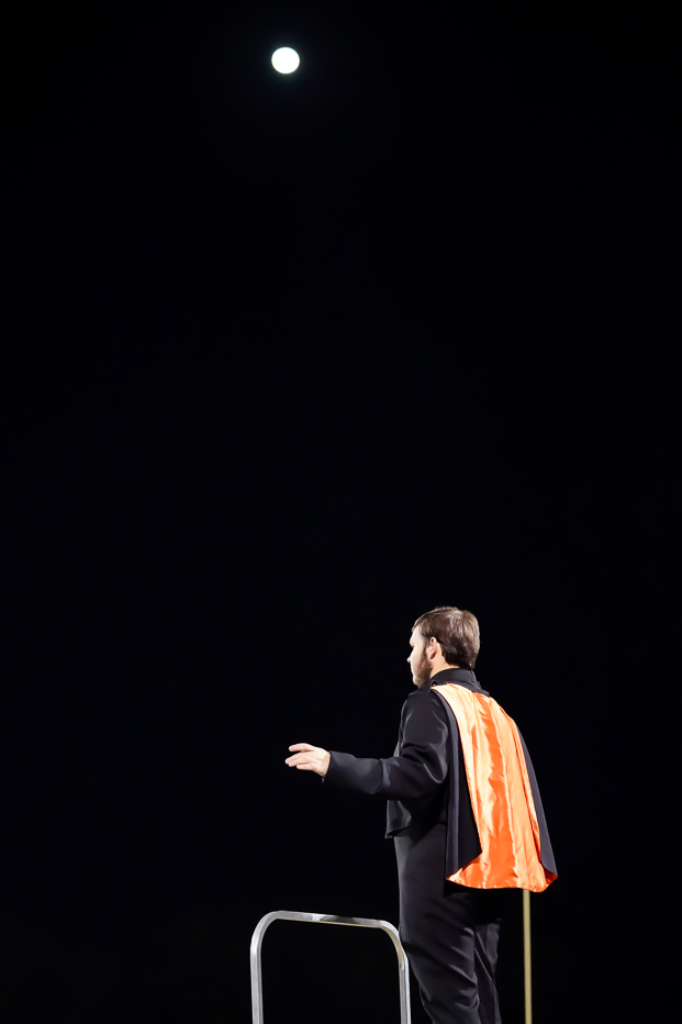 A person wearing a suit and an orange safety vest stands on a platform at night, conducting with their hands raised, beneath a bright full moon in the dark sky.
