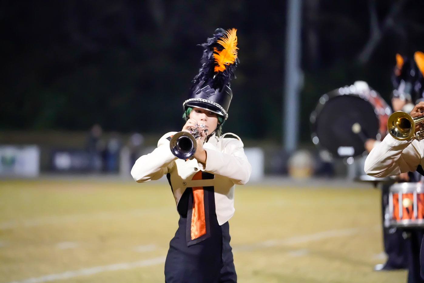 A marching band member in a black and white uniform with an orange and black plume on their hat plays a trumpet on a football field at night. Other band members and a drum are visible in the background.