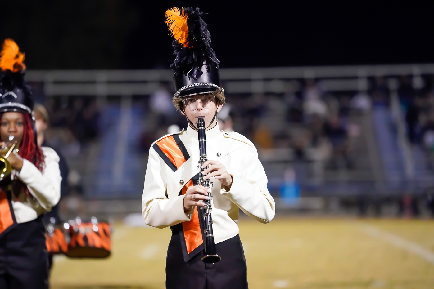 A marching band member in a white and black uniform with orange accents plays a clarinet on a football field at night. Feathers decorate the tall black hat, and blurred spectators are visible in the background.