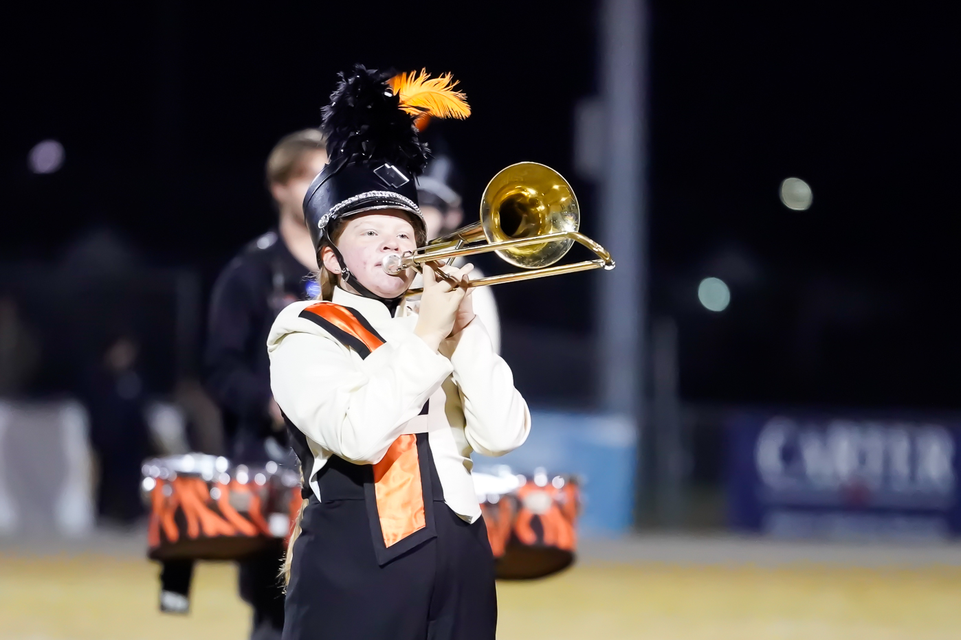 A young marching band member in uniform plays a trombone at night, with another musician blurred in the background holding snare drums. The uniform features a plume-topped hat and an orange sash.