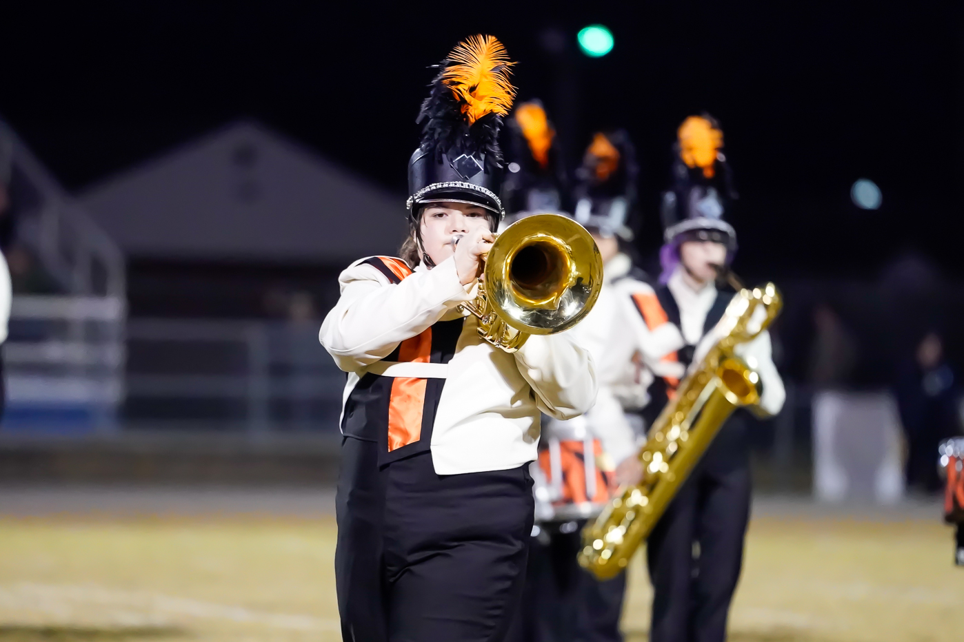 A marching band member in uniform plays a brass instrument on a field at night. Other band members with similar uniforms and hats featuring orange plumes are visible in the background.