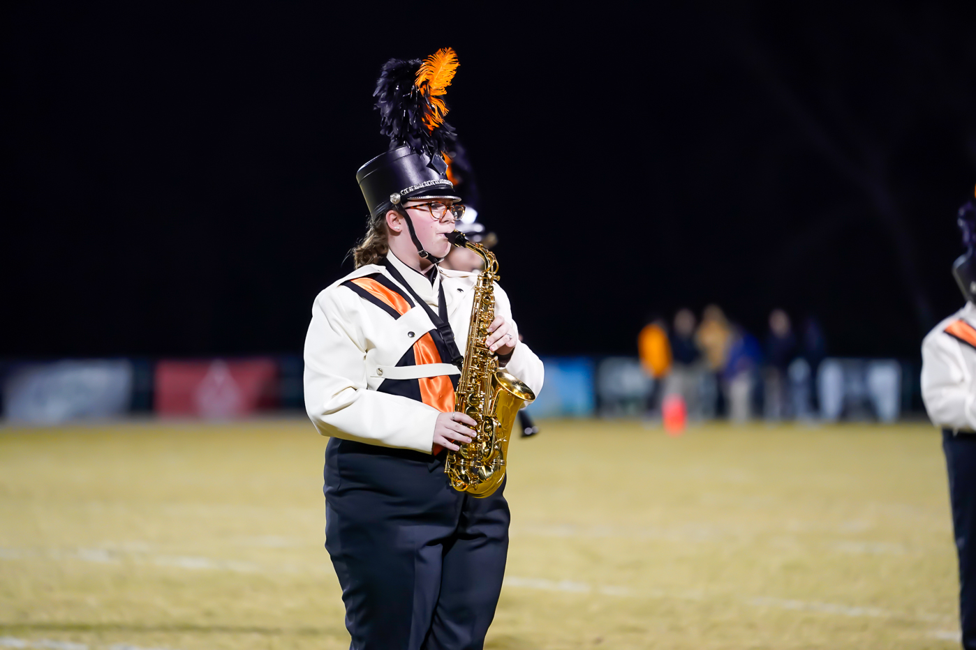 A marching band member in a black and white uniform with orange accents plays a saxophone on a field at night, wearing a feathered hat. Other band members and blurred spectators are visible in the background.