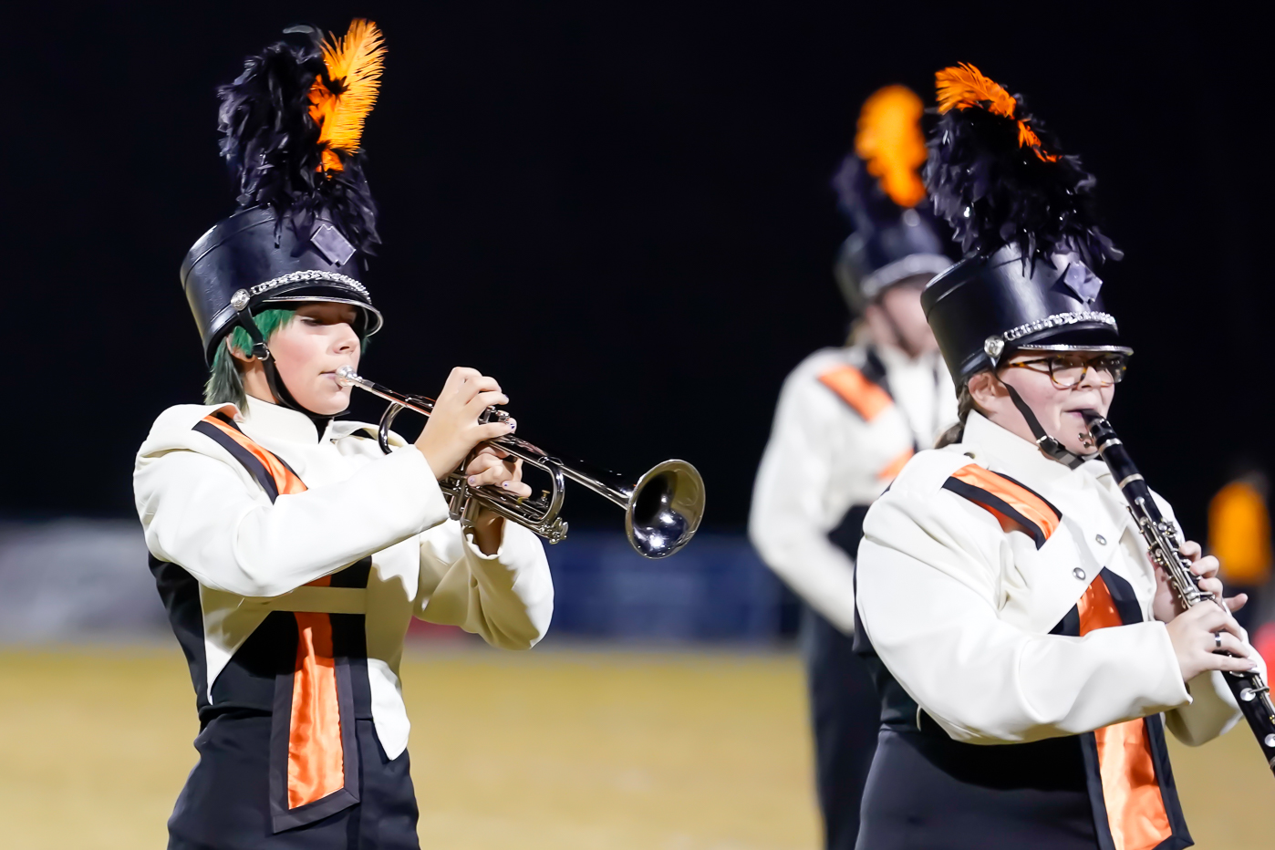 Two marching band members in black and white uniforms with orange accents play brass and woodwind instruments on a field at night. They wear tall hats with black and orange plumes.